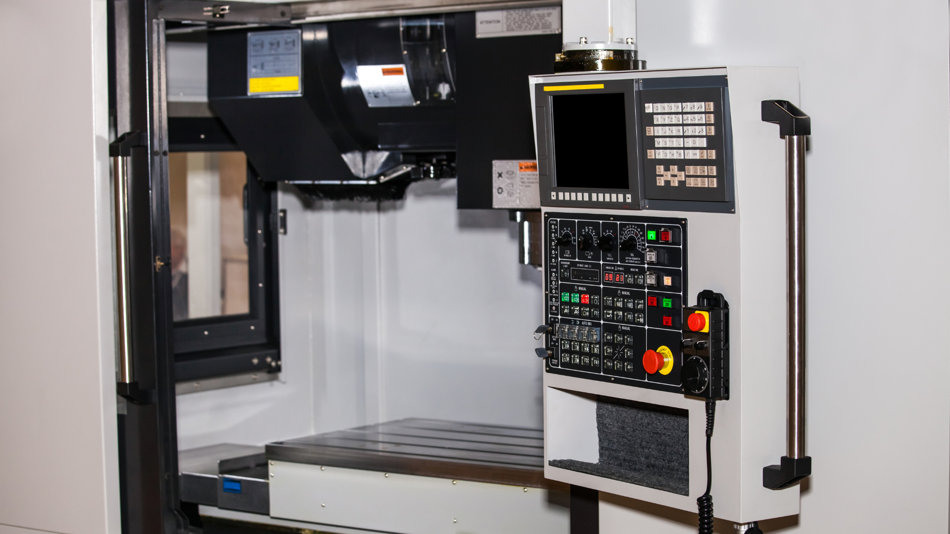 A vertical machining centre with its sliding door open on an empty T-slotted table and spindle head, beside a control console with a pendant handwheel.