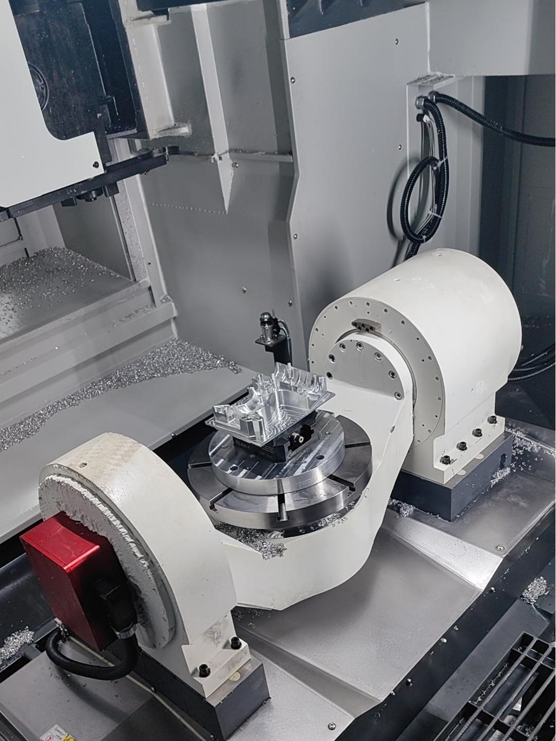 Interior of an enclosed machining centre: a ribbed aluminium part clamped on a trunnion rotary table between two white trunnion housings, with swarf on the machine bed.
