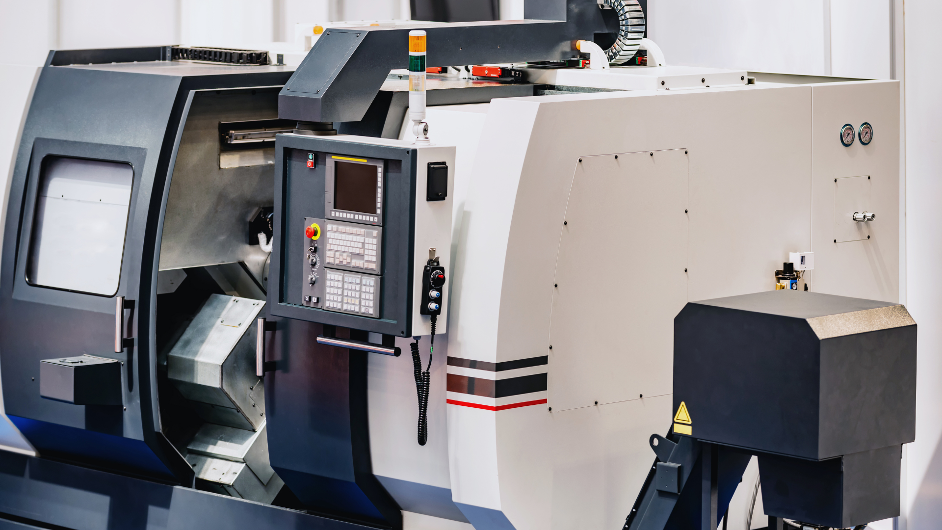 Enclosed CNC turning centre in a machining bay: navy and off-white bodywork, a control console on a swivel arm with a pendant handwheel, doors closed over an empty working area.