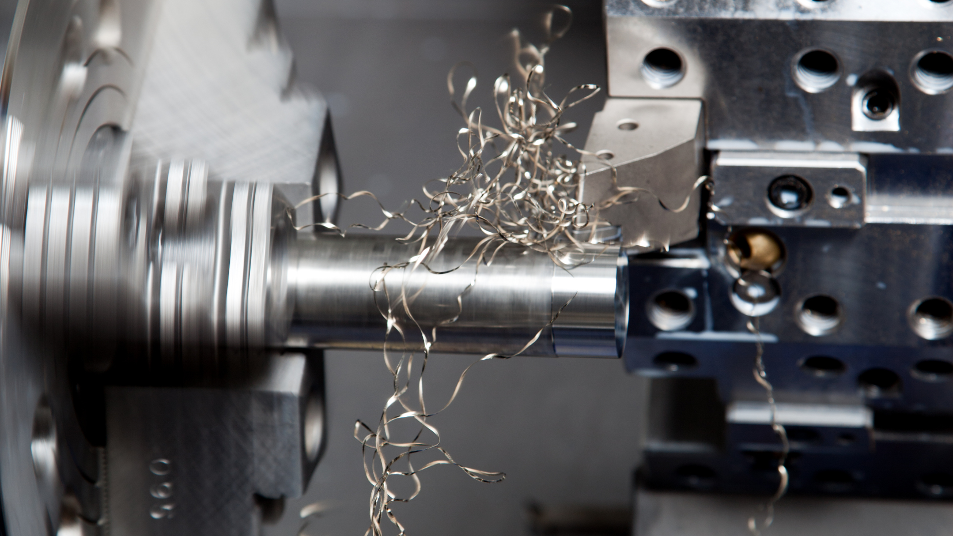 Close view of a turning cut: a cutting tool shearing a rotating workpiece held in the chuck, with curled steel swarf gathered at the tool edge.